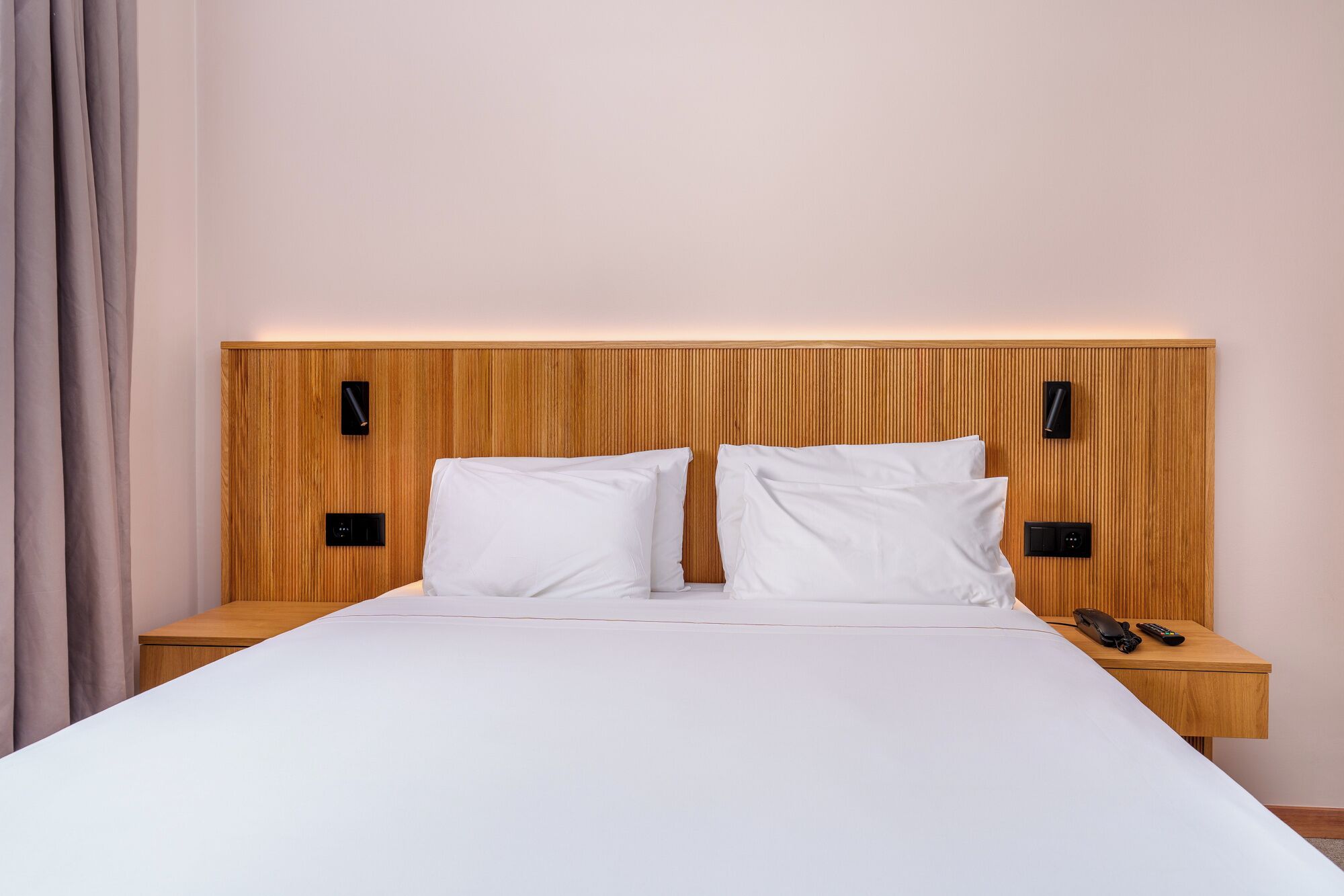 Een hotelkamer met een houten hoofdbord, twee witte kussens, een nachtkastje van hout met een telefoon en afstandsbediening, en een bed met wit beddengoed.