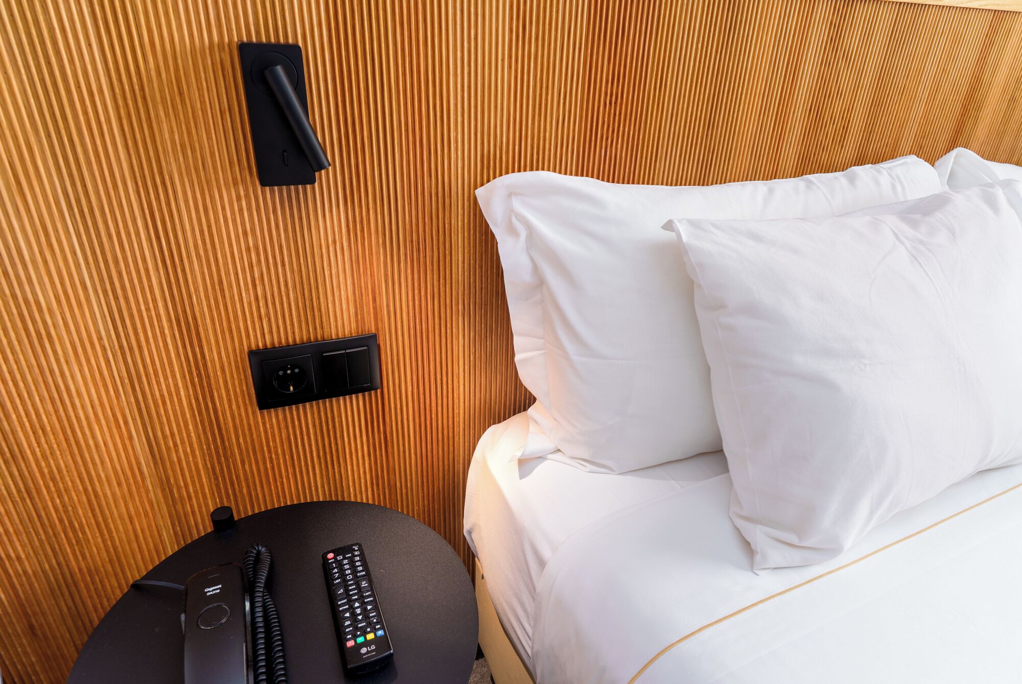 Habitación de hotel con cabecera de madera, cama con ropa blanca, control remoto y toma de corriente.