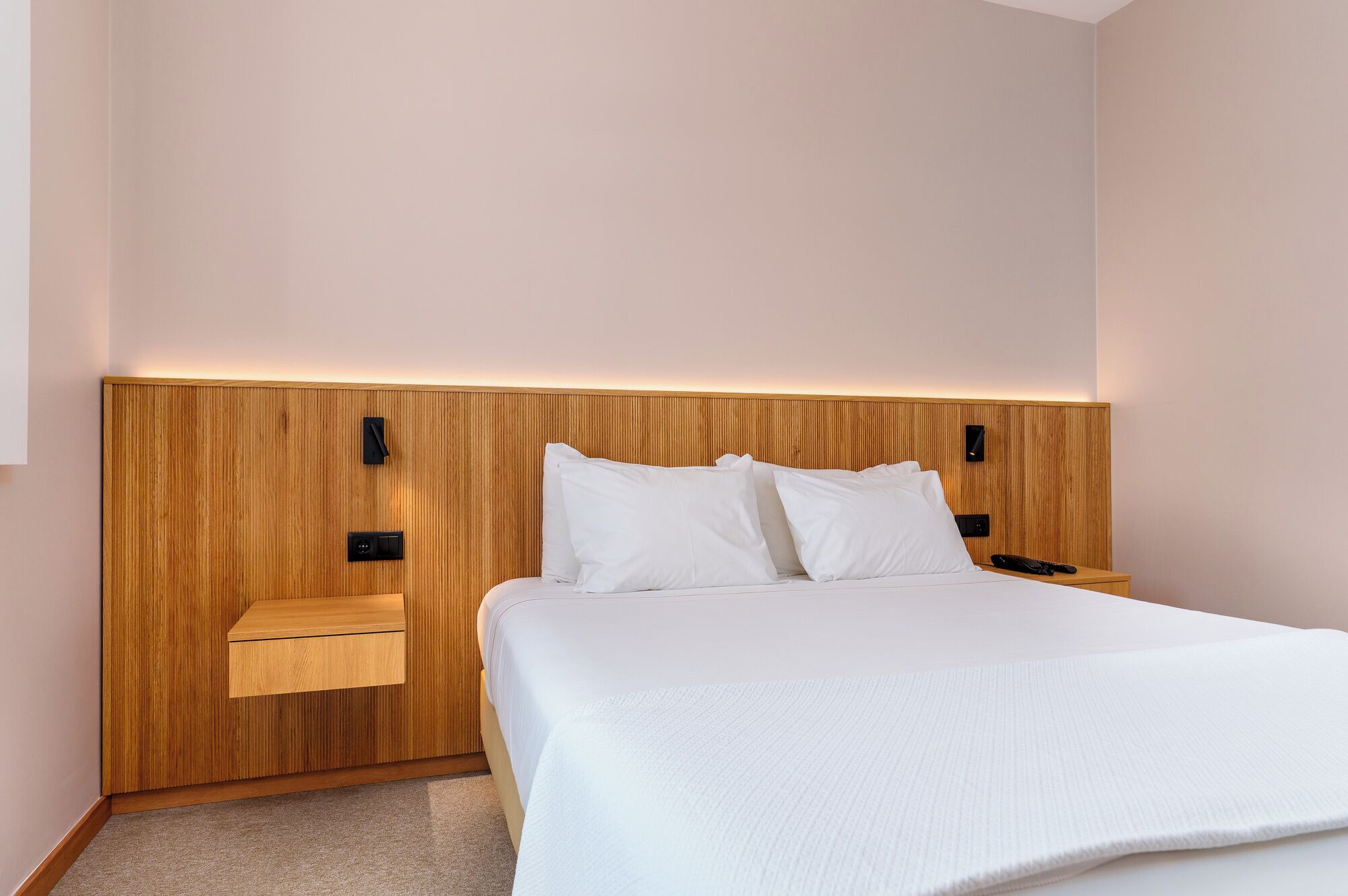 A modern hotel room with a wooden headboard, a large bed, two bedside tables, and a wall-mounted light.