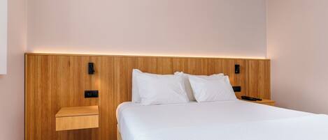 A modern hotel room with a wooden headboard, a large bed, two bedside tables, and a wall-mounted light.