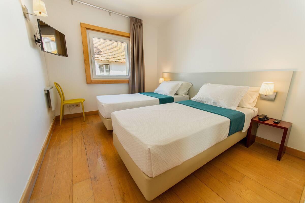 Habitación de hotel con dos camas, una mesita, una silla, ventana con persianas y televisor montado en la pared.