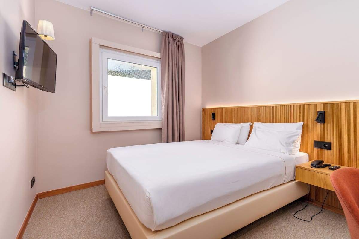 Una habitación de hotel con una cama, televisión montada en la pared, ventana con cortinas, cabecera de madera y mesita de noche con teléfono y control remoto.