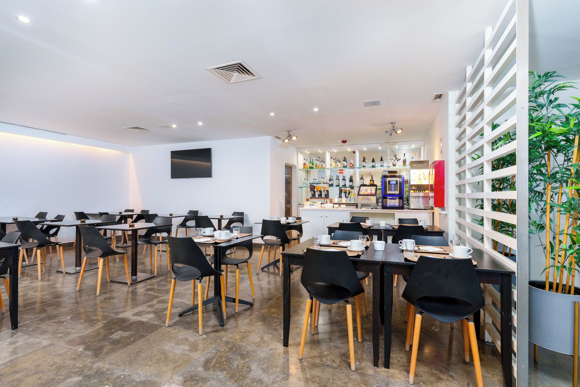A modern dining area with multiple tables and chairs, a bar area with a variety of beverages, and a wall-mounted television.