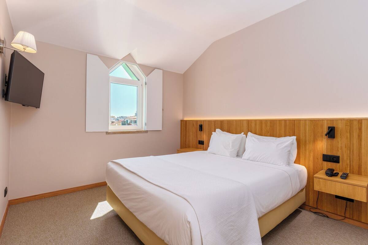 Un dormitorio con cama, mesita de noche, televisor montado en la pared y ventana con vistas.