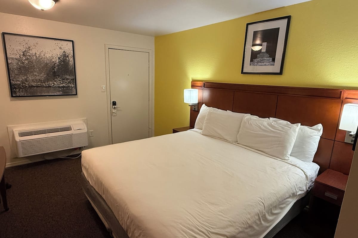 A hotel room with a bed, a wall-mounted air conditioner, a door, and two framed pictures on the wall.