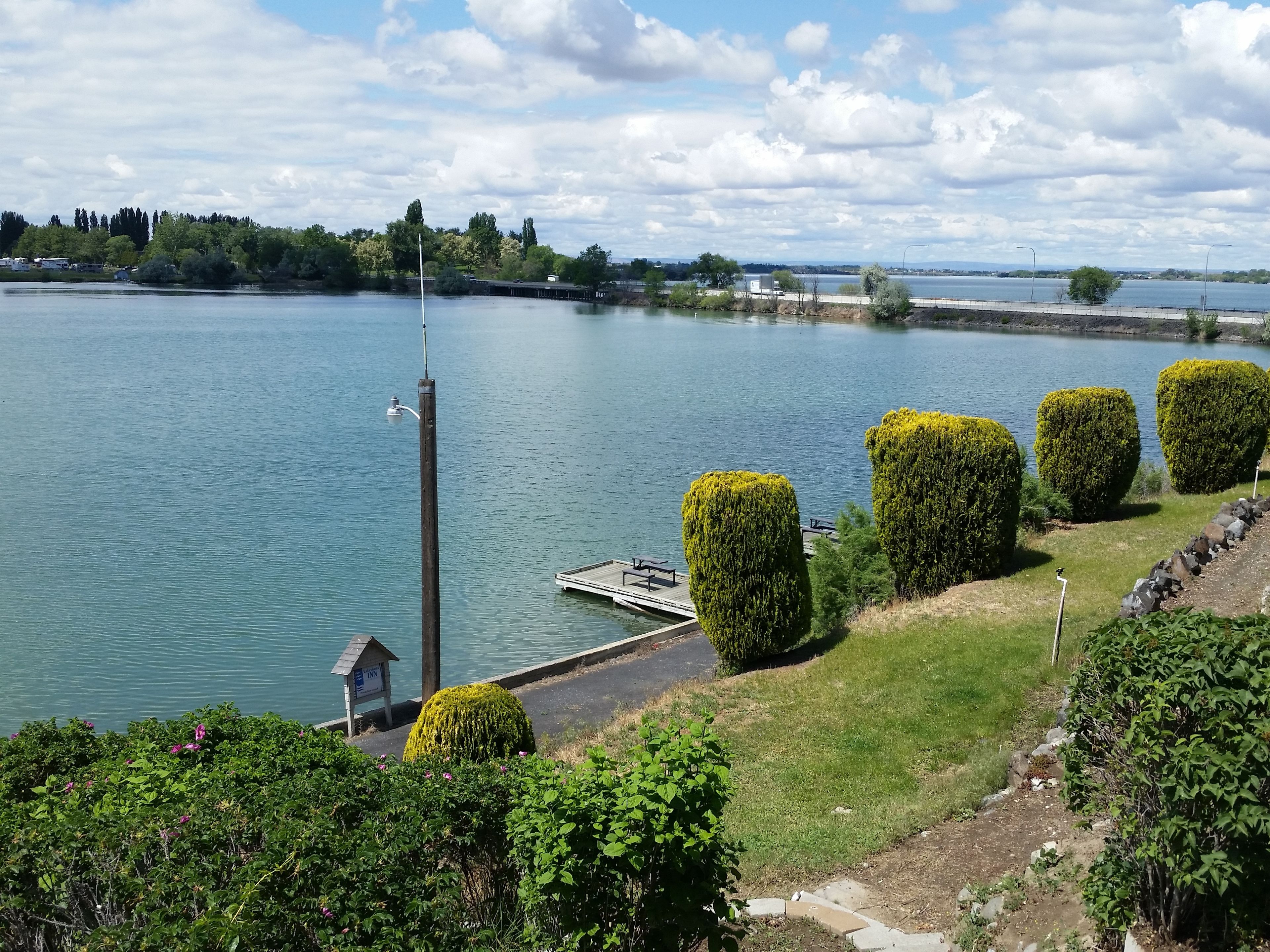 Vista del lago con un muelle, arbustos recortados y una construcción pequeña cerca del agua.