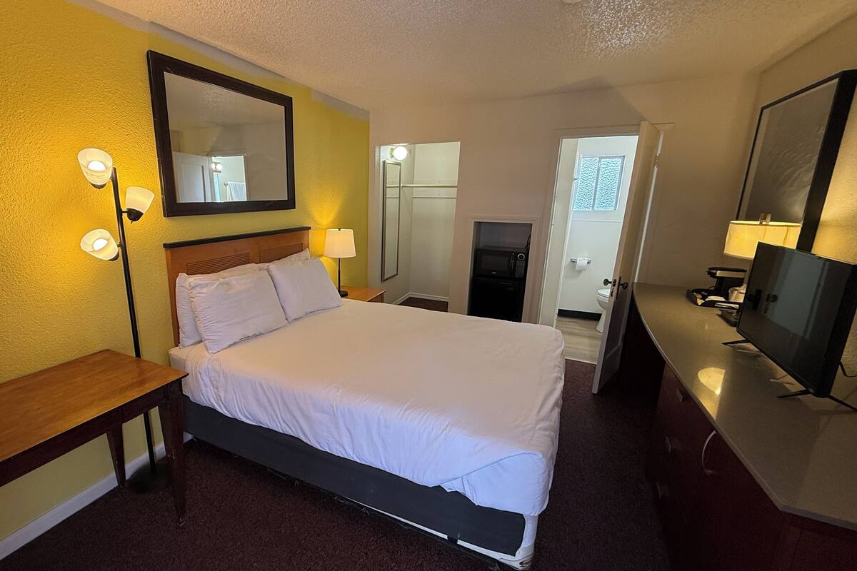 A hotel room with a bed, bedside lamps, a desk, a TV, and a bathroom visible through an open door.