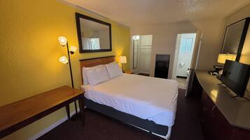 A hotel room with a bed, bedside lamps, a desk, a TV, and a bathroom visible through an open door.