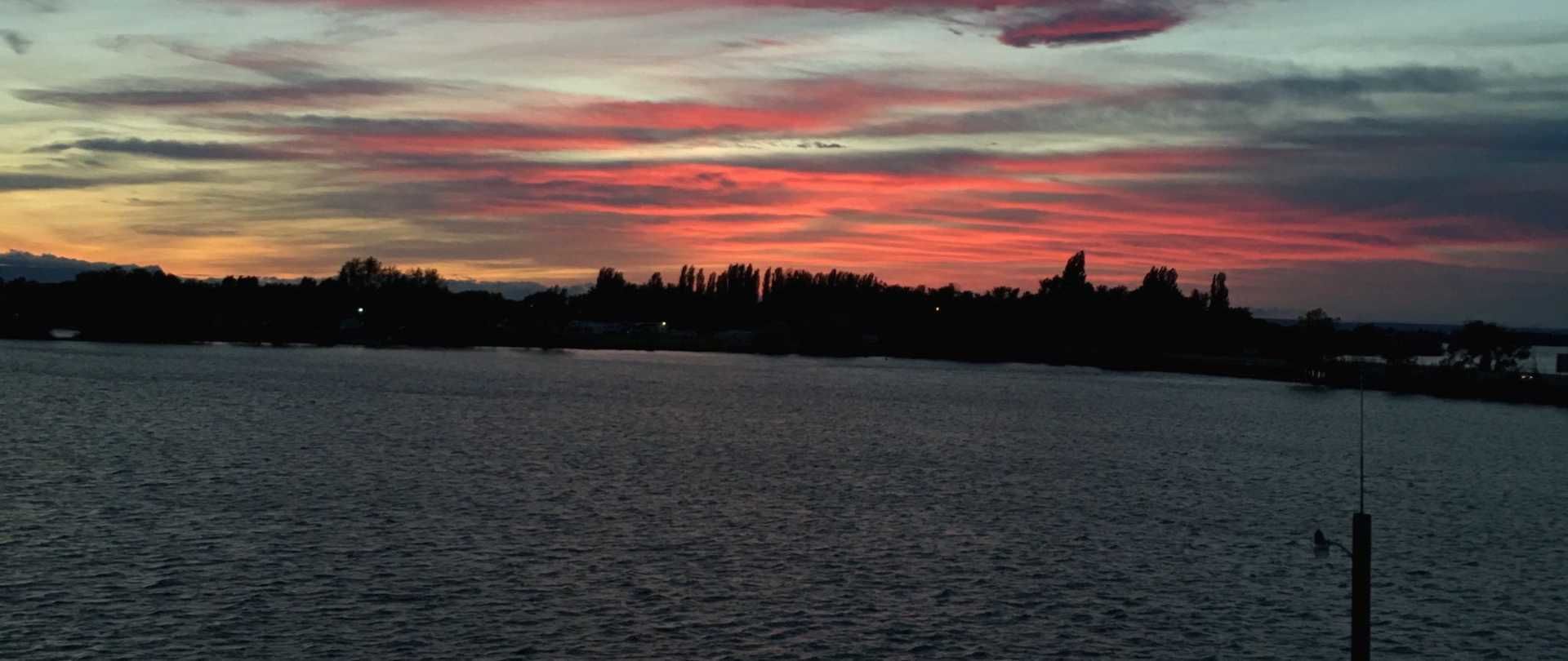 A sunset over a body of water with a silhouette of trees and buildings in the distance.