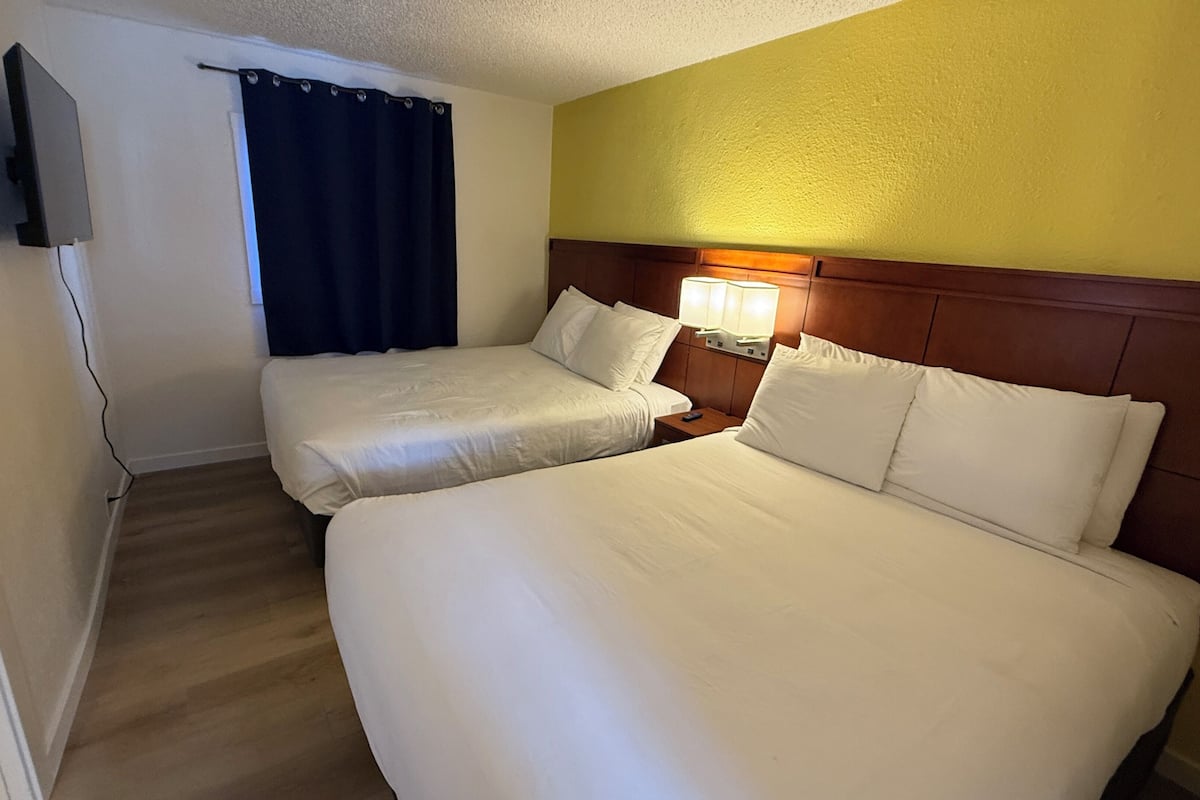 A hotel room with two beds, a TV mounted on the wall, a bedside table with a lamp, and a wooden headboard.