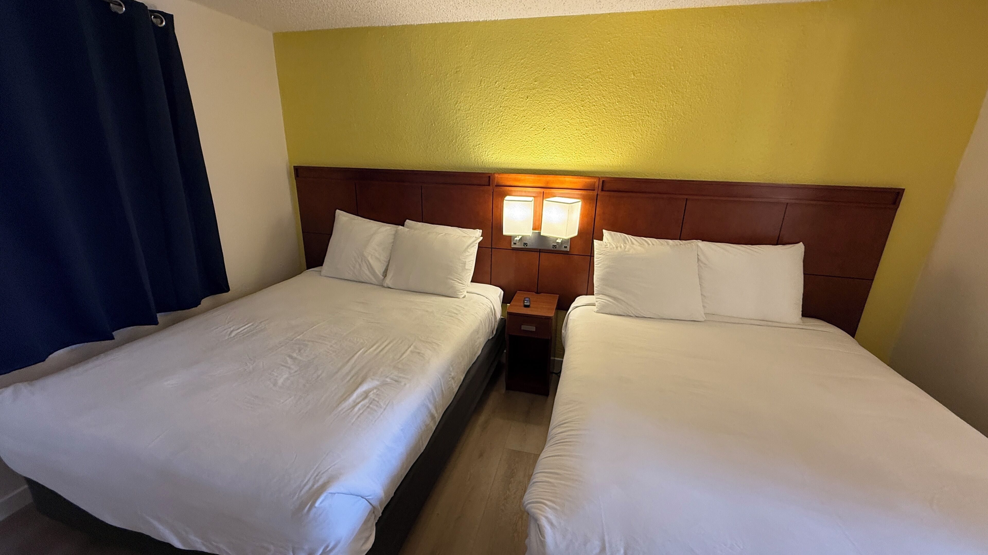Habitación de hotel con dos camas individuales, cabecera de madera, mesita de noche con lámpara y cortina azul.