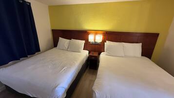 Habitación de hotel con dos camas individuales, cabecera de madera, mesita de noche con lámpara y cortina azul.