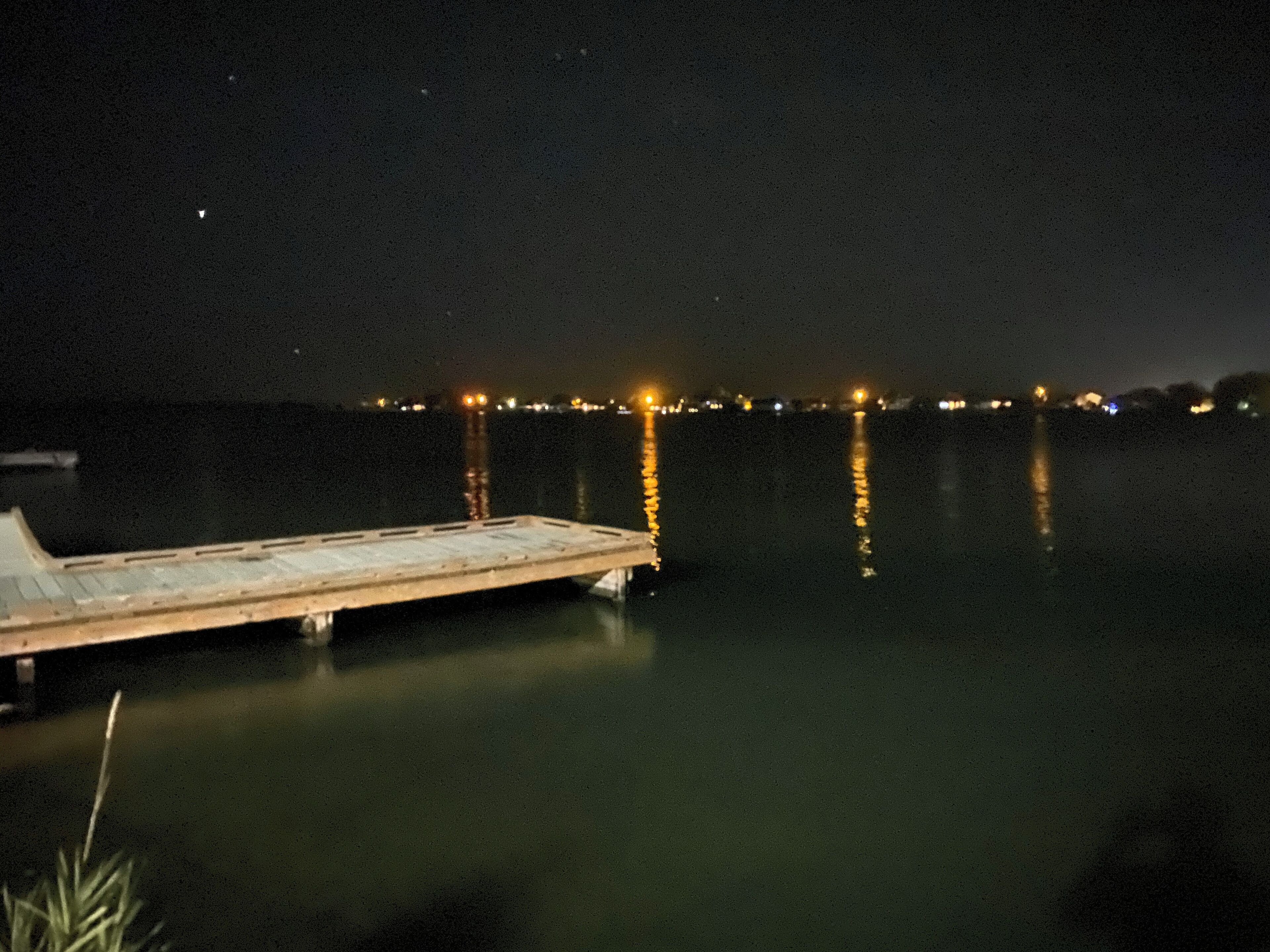 Eine Mole, die sich in ruhiges Wasser erstreckt, bei Nacht mit fernen Stadtlichtern, die sich auf der Oberfläche spiegeln.