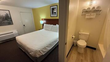 A hotel room with a bed, a heater, a framed picture on the wall, and a bathroom with a toilet and towel rack.