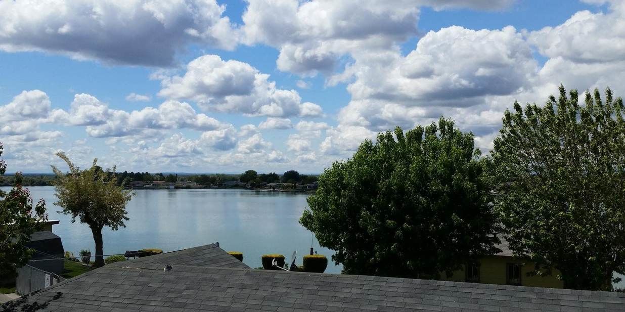 A view of a lake with a clear sky and clouds, surrounded by trees and a building with a sloped roof.