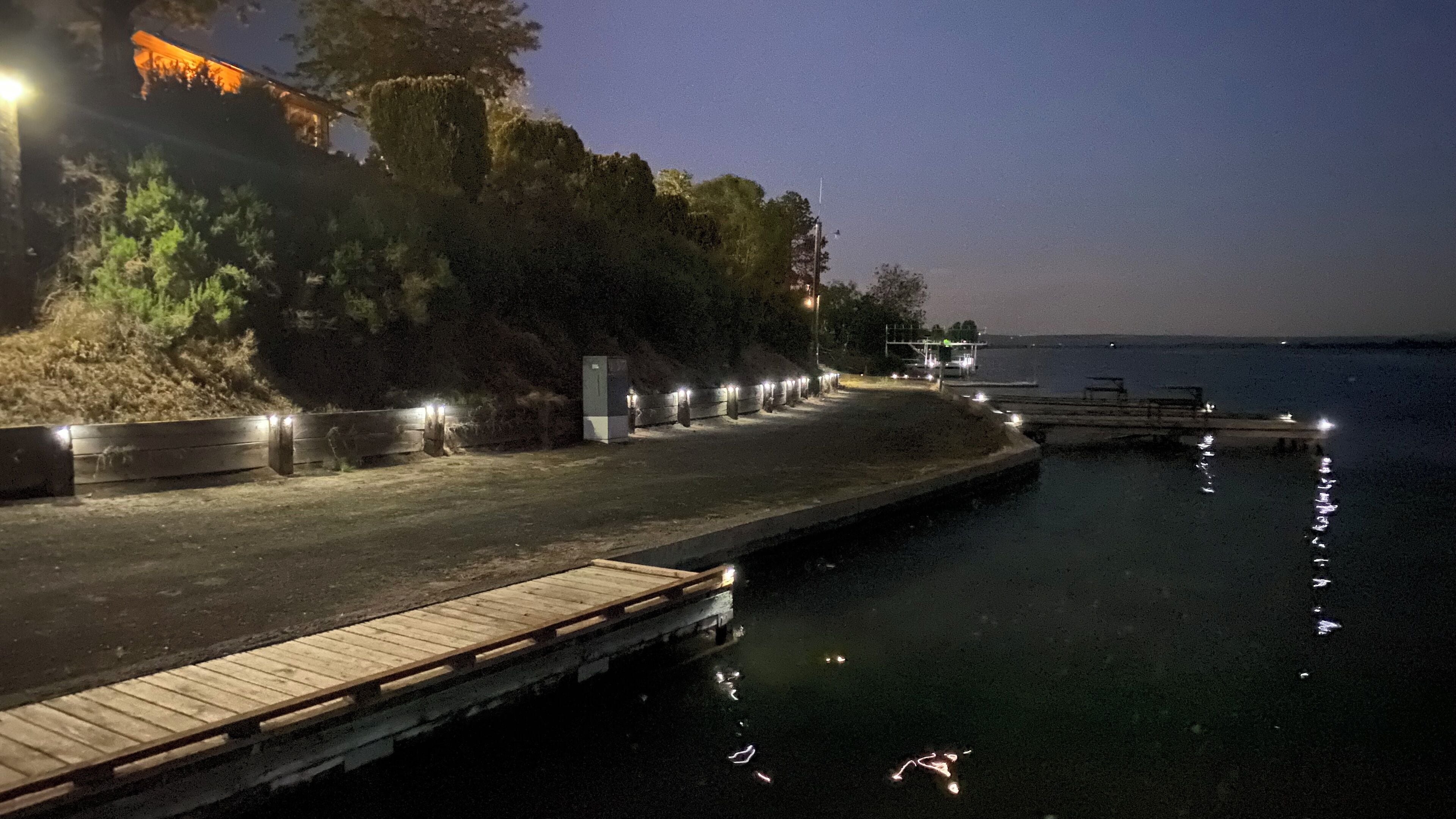 A nighttime waterfront scene with a wooden dock extending into calm water, surrounded by trees and illuminated by streetlights.