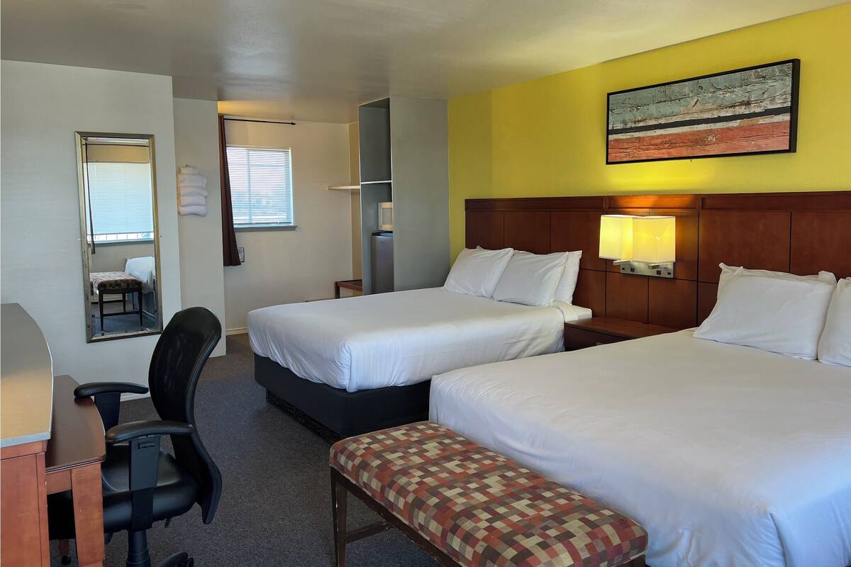 A hotel room with two beds, a desk, a chair, a bench, a mirror, a window with blinds, a wall-mounted TV, and a painting.