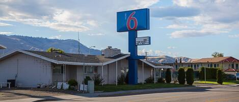 Un motel con un llamativo letrero con el número '6', un estacionamiento y montañas al fondo.