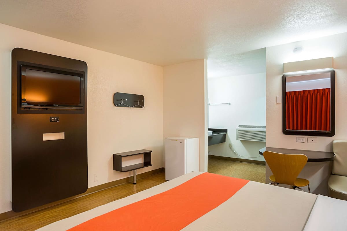A hotel room with a bed, a desk, a chair, a small table, a mirror, a heater, a wardrobe, and a coat rack.