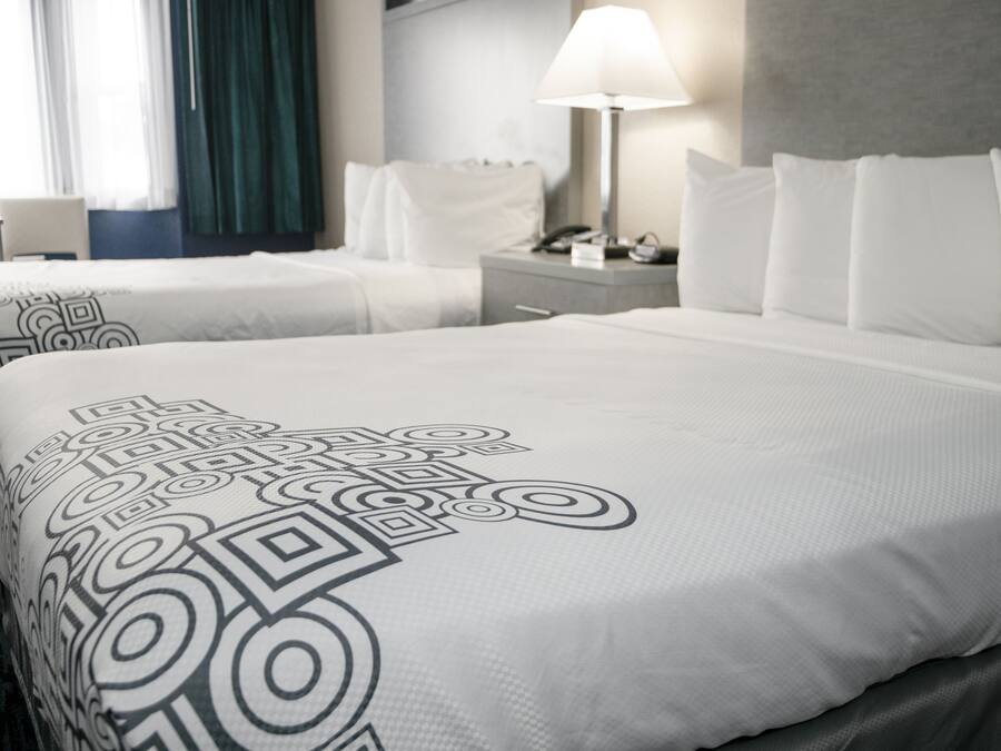 A hotel room with two beds, a nightstand with a lamp, and a view of the window with curtains.