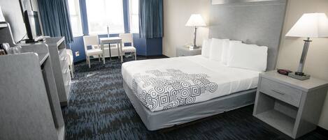 A hotel room with a large bed, two bedside tables with lamps, a desk with a chair, a TV, and a window with curtains.