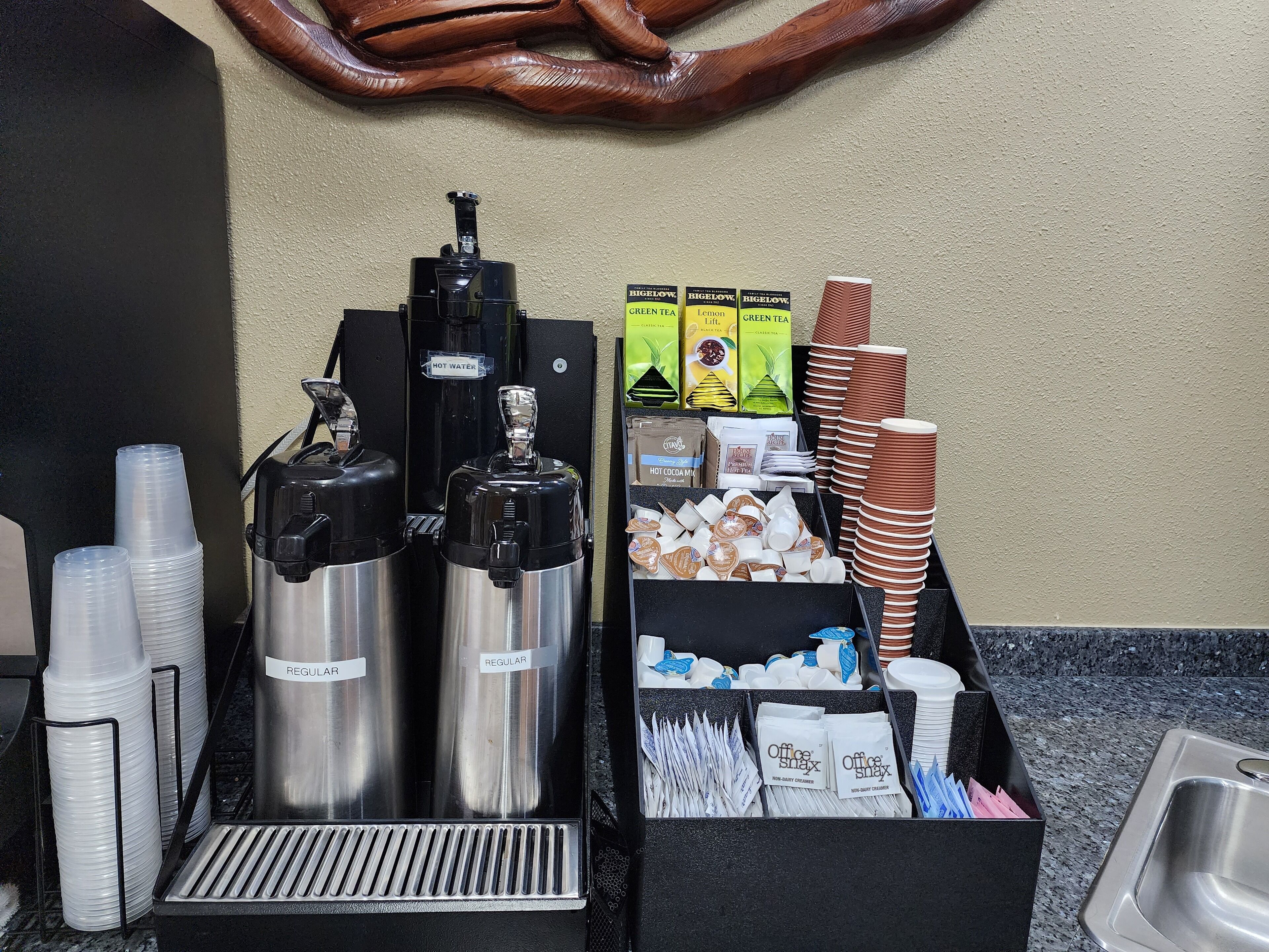A coffee station with two hot water dispensers, a variety of coffee pods, disposable cups, and a sink.