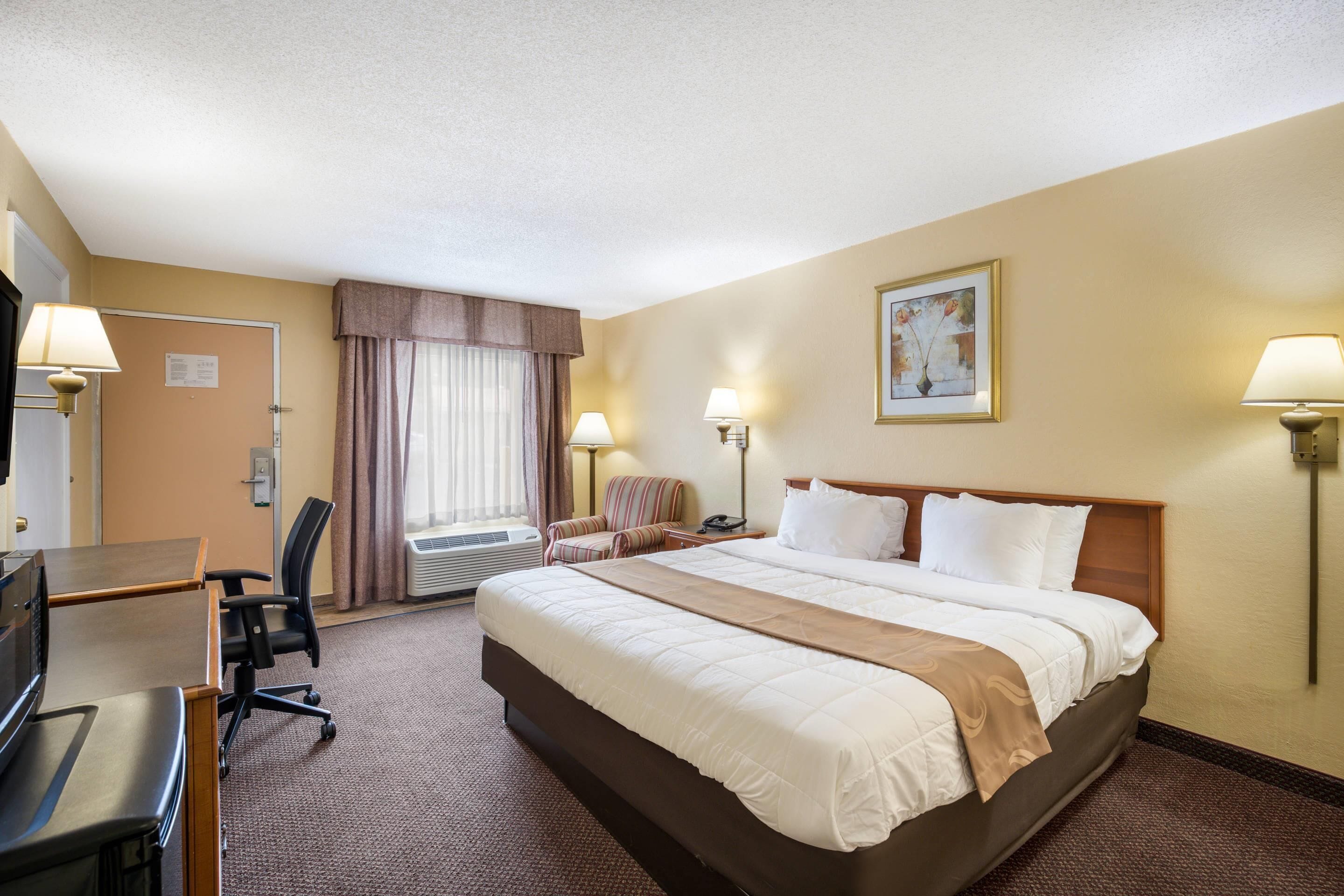A hotel room with a large bed, a desk, a chair, a desk lamp, a wall-mounted lamp, a painting, and a window with curtains.