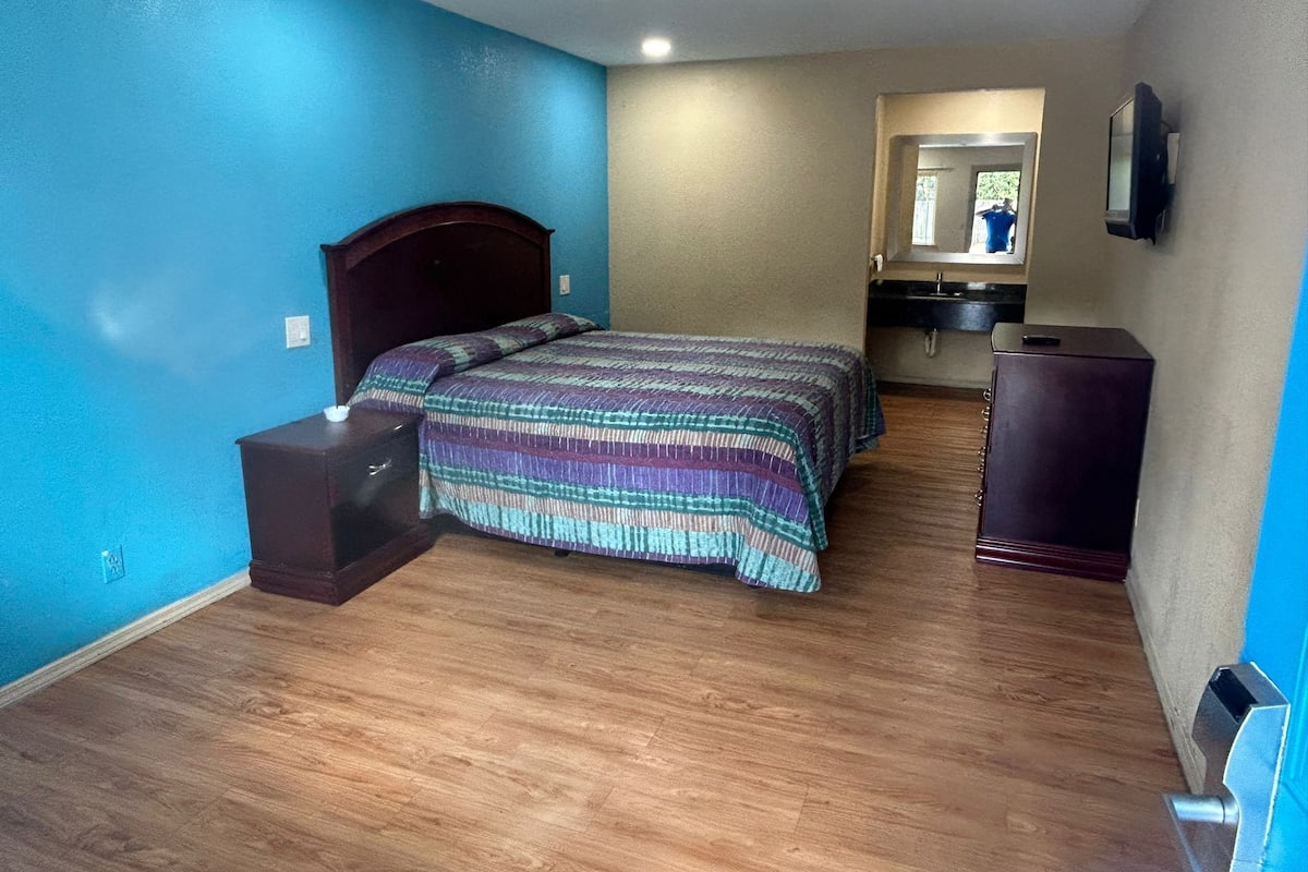 Un dormitorio con cama, piso de madera, cómoda, espejo y televisor.