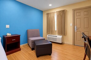 A hotel room with a blue wall, a grey sofa, a wooden floor, a small desk with a telephone, a window with curtains, and a door with a handle. - Blue Star Inn Lafayette Near Airport I-10 (Lafayette)