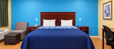 A hotel room with a large bed, a desk, a chair, a nightstand, and a wall-mounted heater.