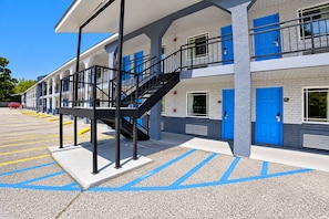 A multi-story hotel with a central staircase, blue doors, and a parking lot with marked spaces. - Blue Star Inn Lafayette Near Airport I-10 (Lafayette)