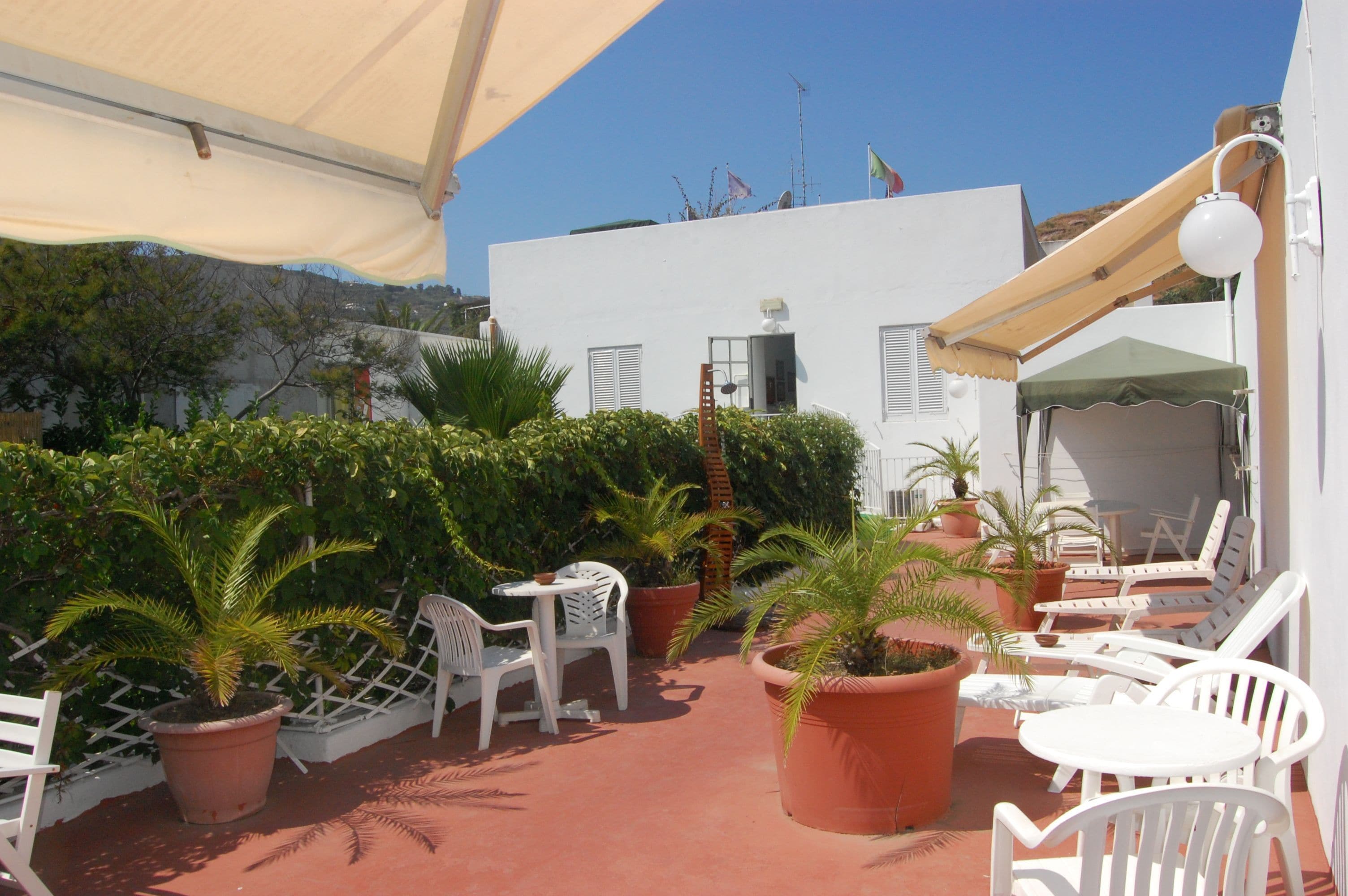 Eine Terrasse mit weißen Stühlen und Tischen, umgeben von Topfpflanzen und einem Gebäude mit rotem Dach.