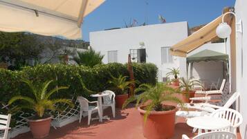 Eine Terrasse mit weißen Stühlen und Tischen, umgeben von Topfpflanzen und einem Gebäude mit rotem Dach.