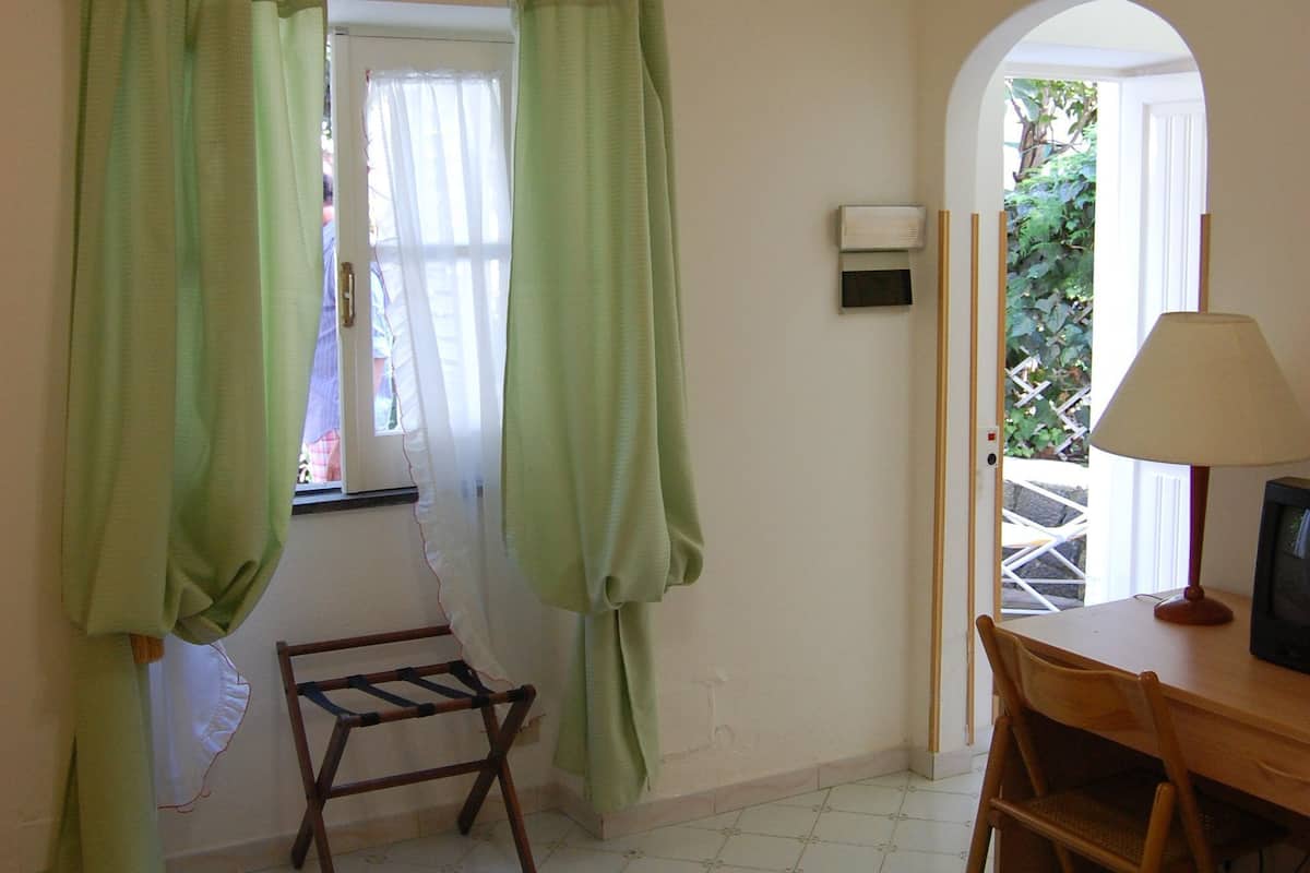 Ein Zimmer mit einem Bett, einem Holzstuhl, einem Tisch mit Lampe, einem Fenster mit Vorhängen und einer Klimaanlage.