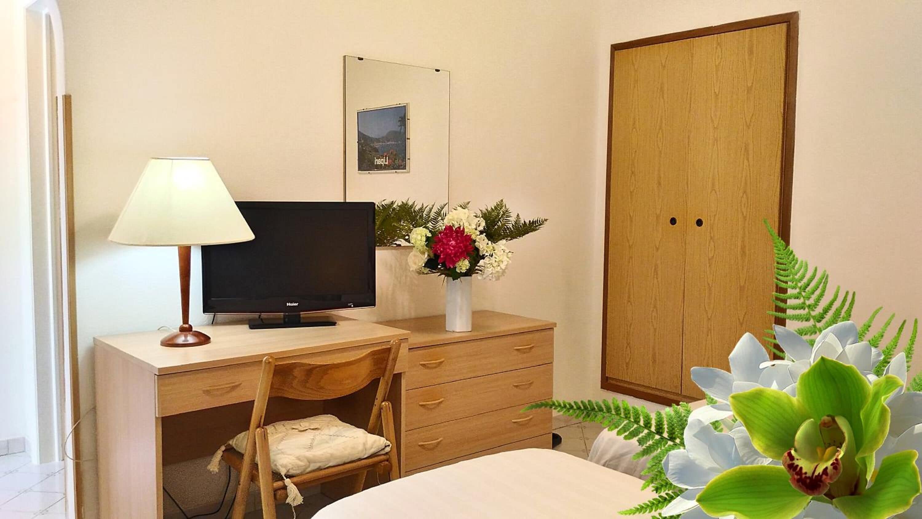 Una stanza con una scrivania, una televisione, una sedia, un armadio e un vaso con fiori.