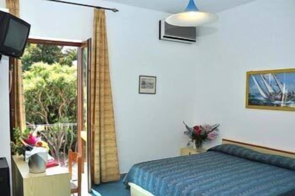 A bedroom with a bed, a TV, a window with curtains, a painting, and a small table with a vase.