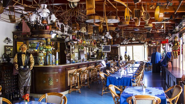 A restaurant with long tables covered in blue tablecloths, wicker chairs, and a bar area with various bottles and decorations. - Hotel Oriente (Lipari)