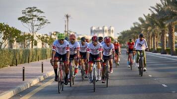 A group of cyclists on a road with palm trees and a building in the background.