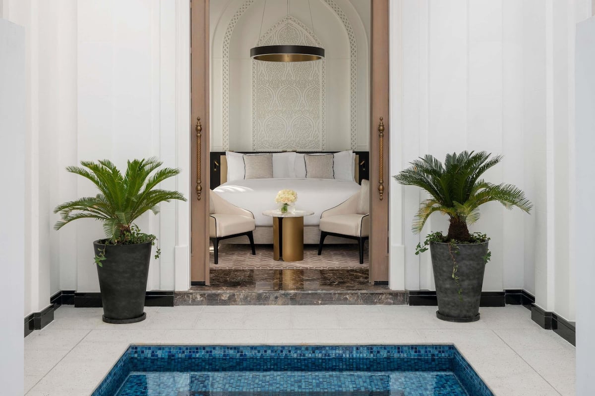 Habitación de hotel con cama, una mesita redonda, dos plantas en macetas y vistas a una piscina.