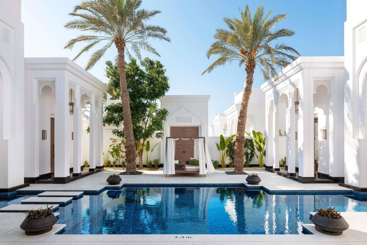 A pool surrounded by white buildings with palm trees, a door, and a small garden area.