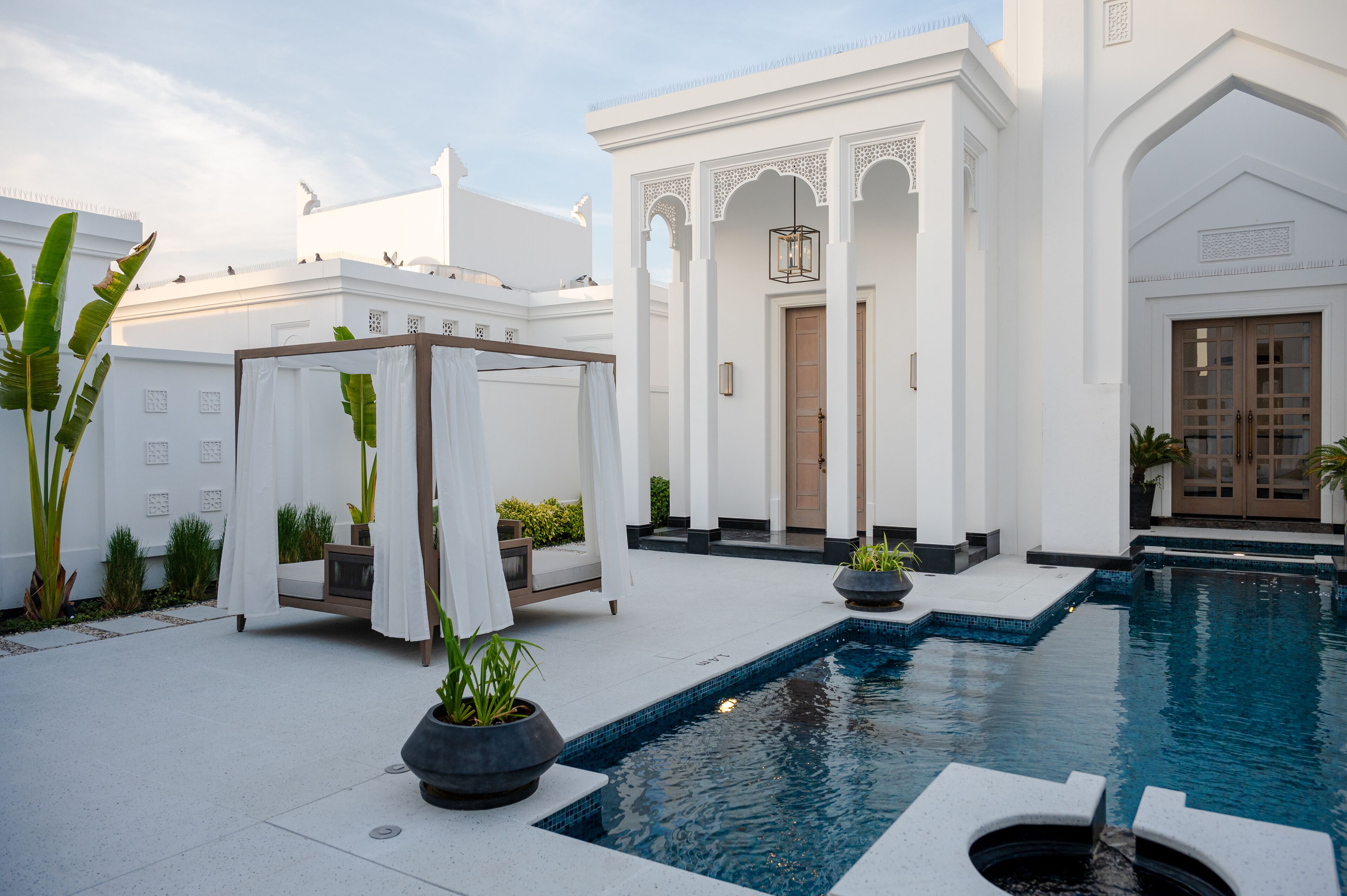 A pool area with a white building, a covered seating area, and potted plants.