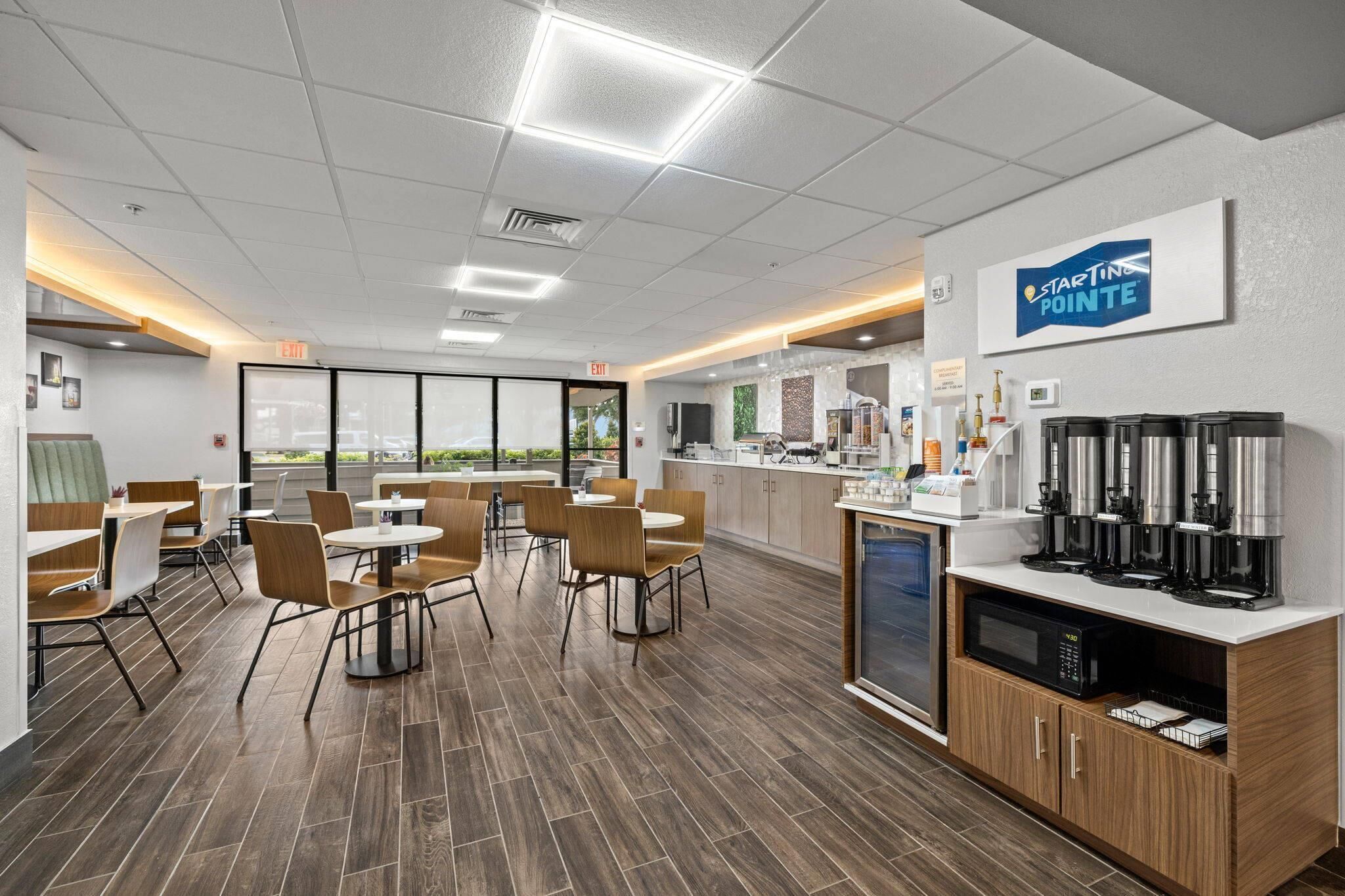 A modern dining area with wooden flooring, round tables, and chairs. There is a coffee station with multiple coffee makers and a microwave. The room has large windows and a view of the outdoors.