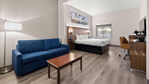 A modern hotel room with a blue sofa, a wooden coffee table, a bed with a headboard, a desk with a computer, and a large window. - Clarion Pointe Lakeland I-4 (Lakeland)