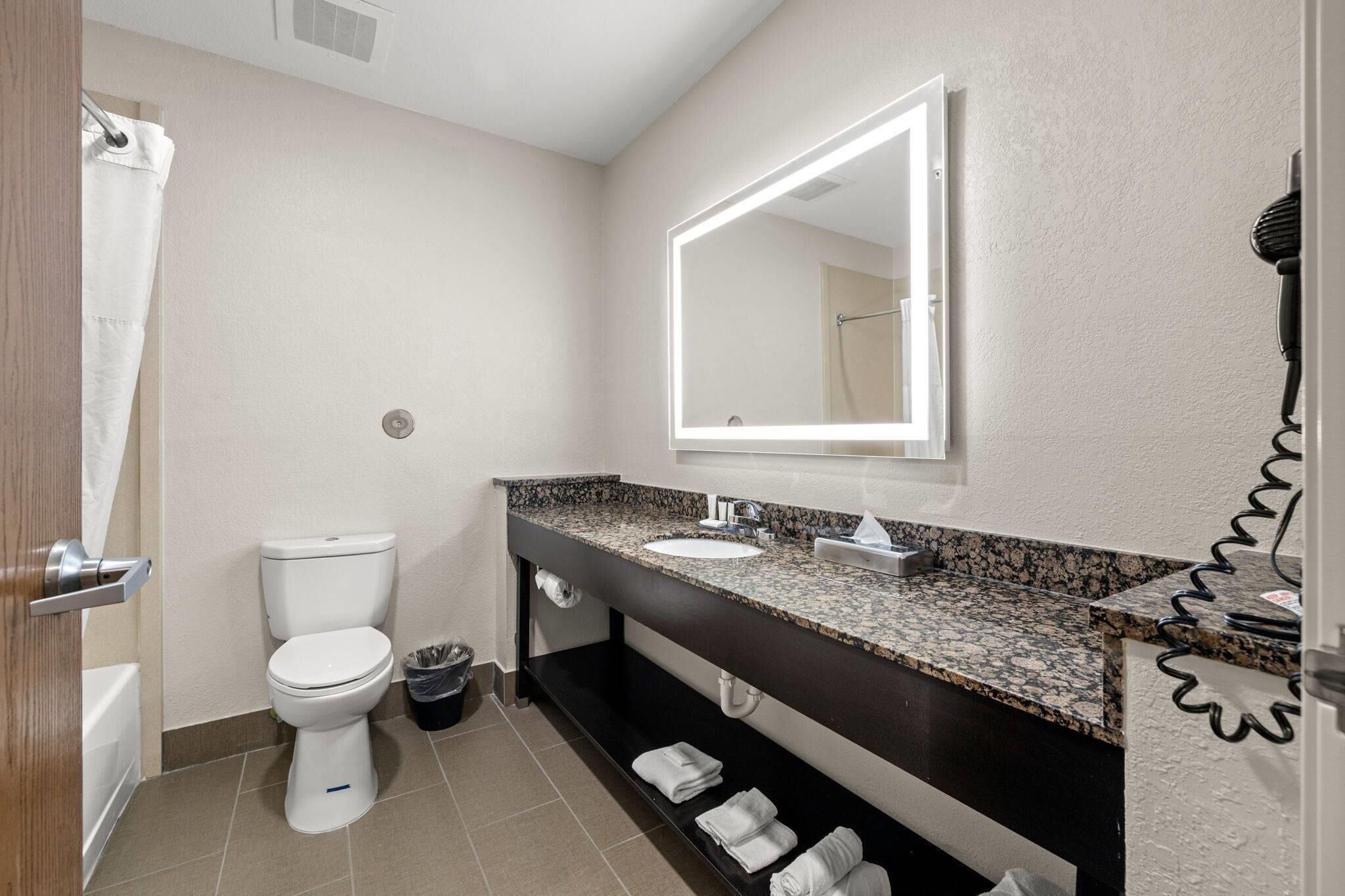 Un baño moderno con inodoro blanco, lavamanos con encimera de granito, un espejo grande y un secador de pelo sobre un soporte.