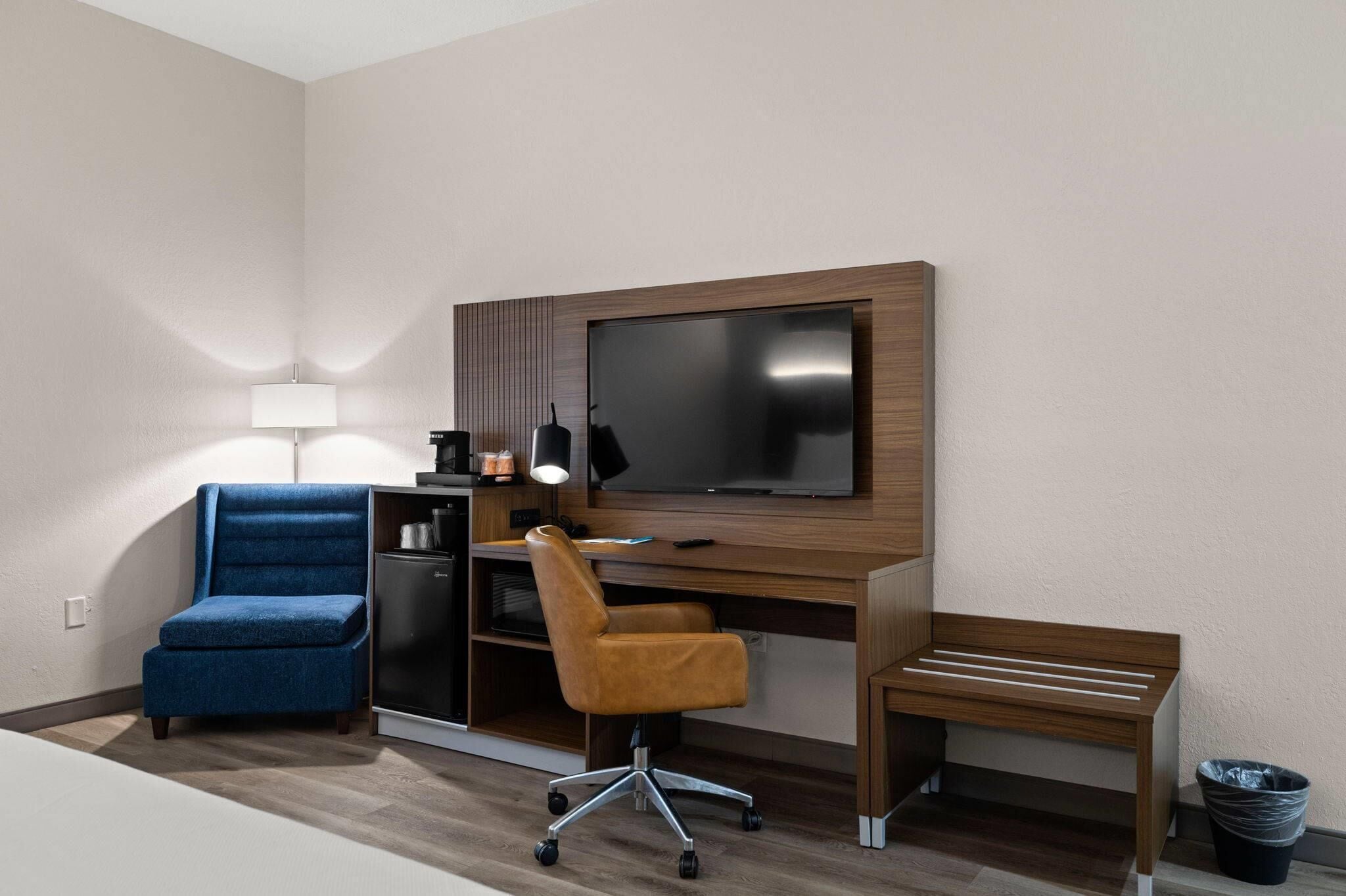 Una habitación de hotel moderna con una televisión de pantalla plana, un escritorio con una computadora, una silla azul, una silla marrón y un pequeño banco.