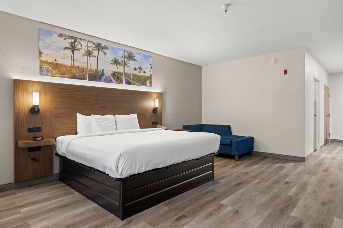 A modern hotel room with a large bed, a wooden headboard, a nightstand, a blue armchair, and a painting of a beach scene.