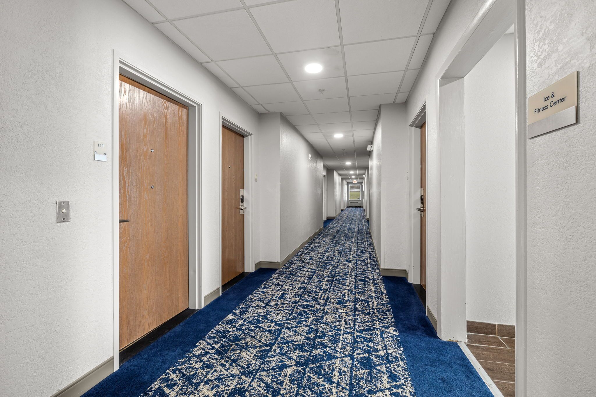 Corredor de hotel com carpete azul, portas de madeira e placa indicando o Centro de Gelo e Fitness.