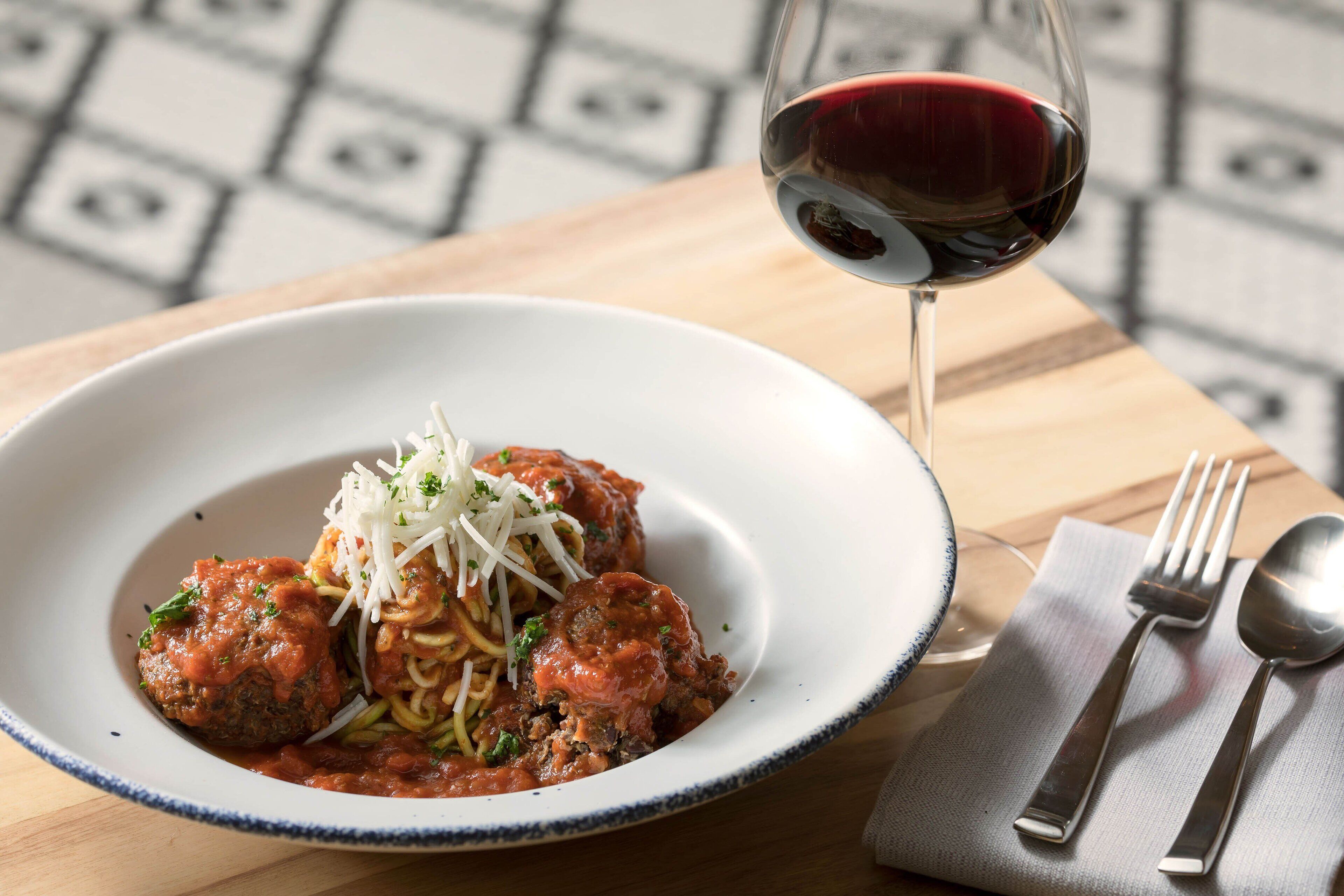 A plate of meatballs with spaghetti and grated cheese, a glass of red wine, and cutlery on a wooden table.