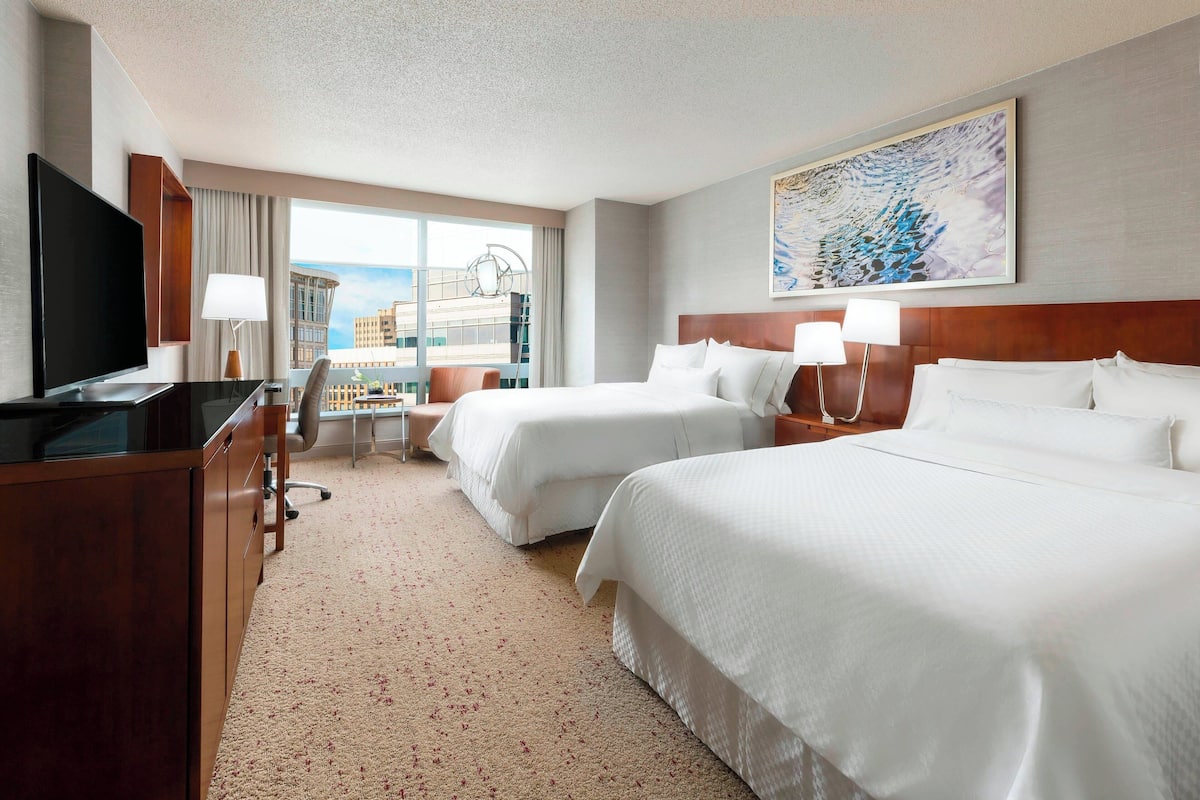 Quarto de hotel com duas camas, uma escrivaninha, televisão e uma janela ampla com vista para edifícios.