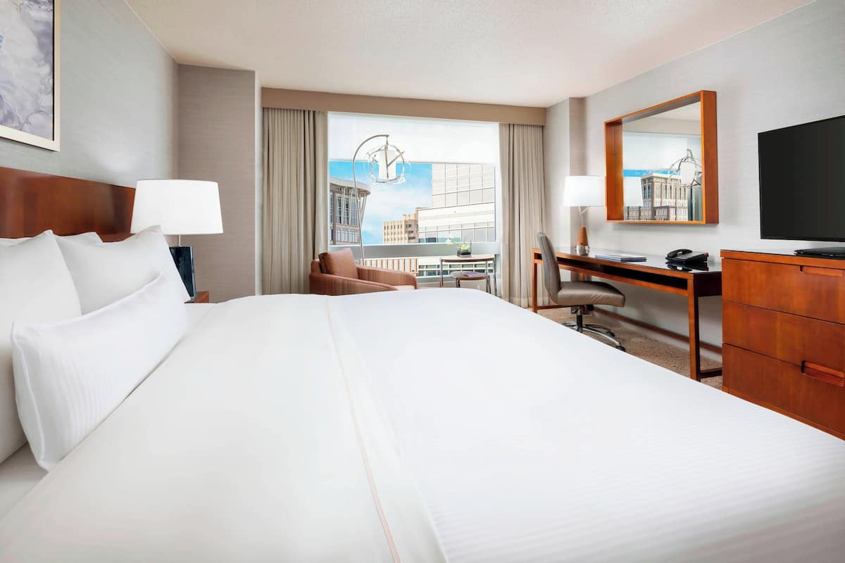 Quarto de hotel com uma cama grande, uma escrivaninha com cadeira, televisão e vista para a paisagem urbana pela janela.